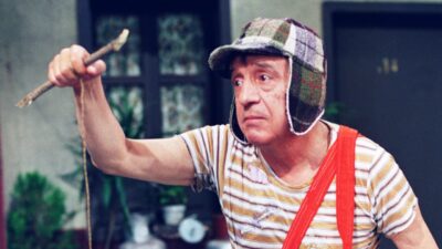 Chaves interpretado por Roberto Gómez Bolaños segura estilingue e levanta objeto em cena no pátio da vila do Chaves