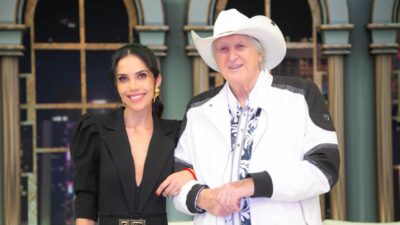 Daniela Albuquerque e Sérgio Reis posam sorrindo lado a lado no palco do Sensacional com cenário de janelas ao fundo