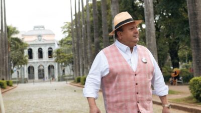Erick Jacquin caminha em rua arborizada usando chapéu e colete claro com construção histórica ao fundo