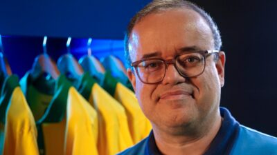 Everaldo Marques sorri usando óculos diante de camisas da seleção brasileira penduradas com iluminação azul ao fundo