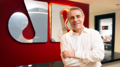 Fernando Pelegio posa de braços cruzados em escritório com parede vermelha e logotipo da Jovem Pan ao fundo