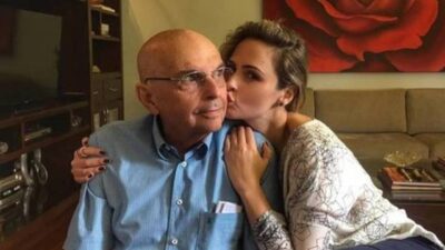 Geraldo Renault sentado em sofá recebe beijo de Ana Paula Renault em sala de estar com quadro de rosa ao fundo