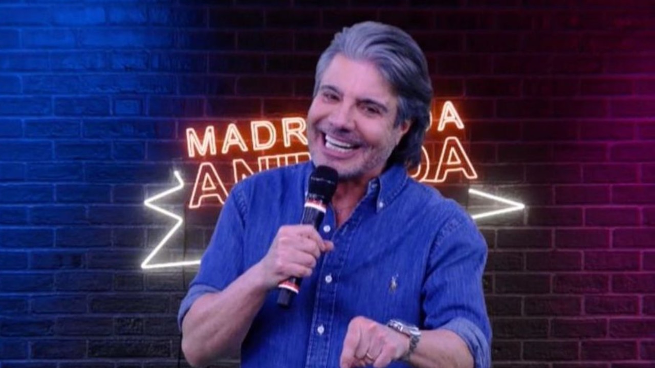 João Kleber segura microfone e sorri em palco com letreiro luminoso ao fundo em cenário de programa de TV