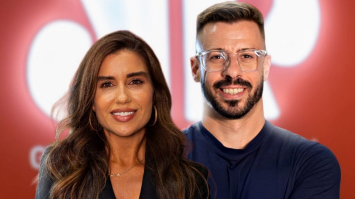 Julia Ballarini e Felippe Facincani posam sorridentes para foto em plano médio, a jornalista veste blazer preto e o comentarista usa camisa azul e óculos de grau em frente a fundo vermelho desfocado