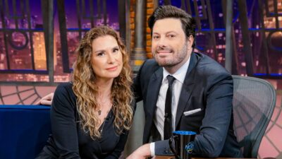 Danilo Gentili e Lavínia Vlasak sentados no cenário do The Noite com caneca do programa e cenário noturno ao fundo