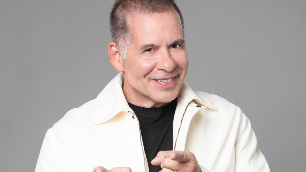 Leandro Hassum sorri e aponta para frente em foto promocional com fundo neutro