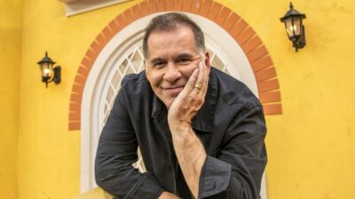 Leandro Hassum apoia o rosto na mão e sorri diante de parede amarela com janela em arco e luminárias externas ao fundo