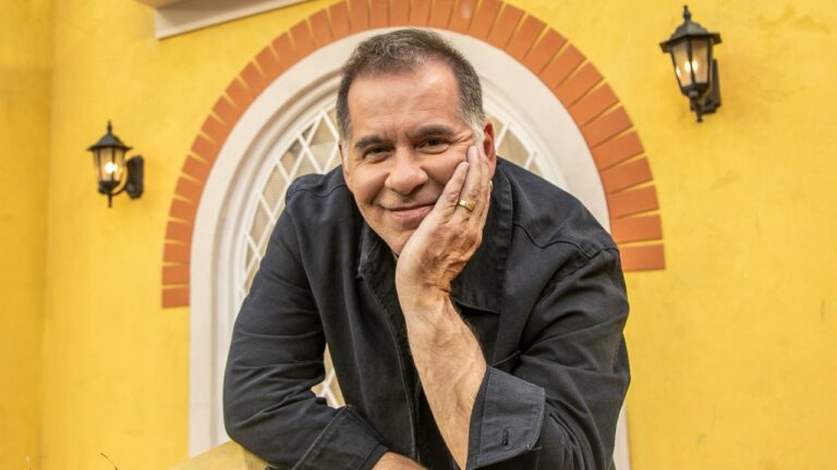 Leandro Hassum apoia o rosto na mão e sorri diante de parede amarela com janela em arco e luminárias externas ao fundo