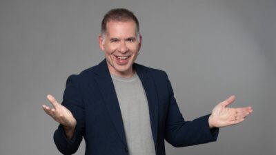 Leandro Hassum sorri com braços abertos usando blazer escuro e camiseta clara diante de fundo neutro em foto de divulgação