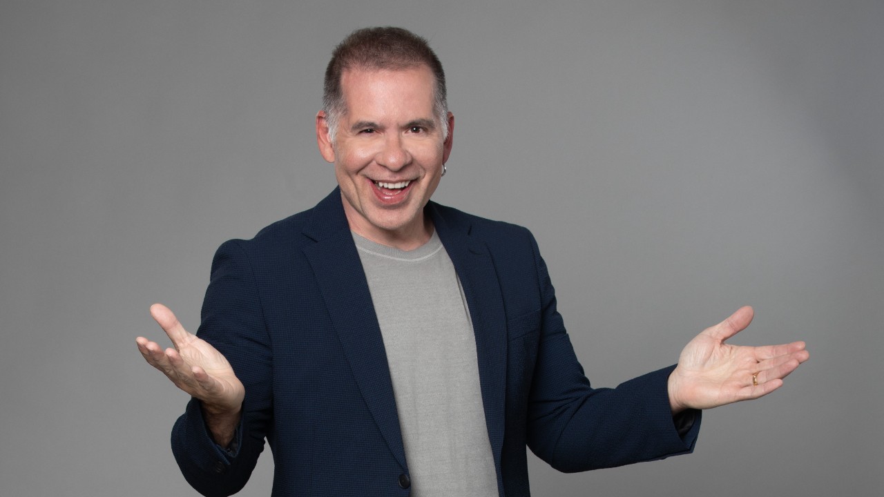 Leandro Hassum sorri com braços abertos usando blazer escuro e camiseta clara diante de fundo neutro em foto de divulgação