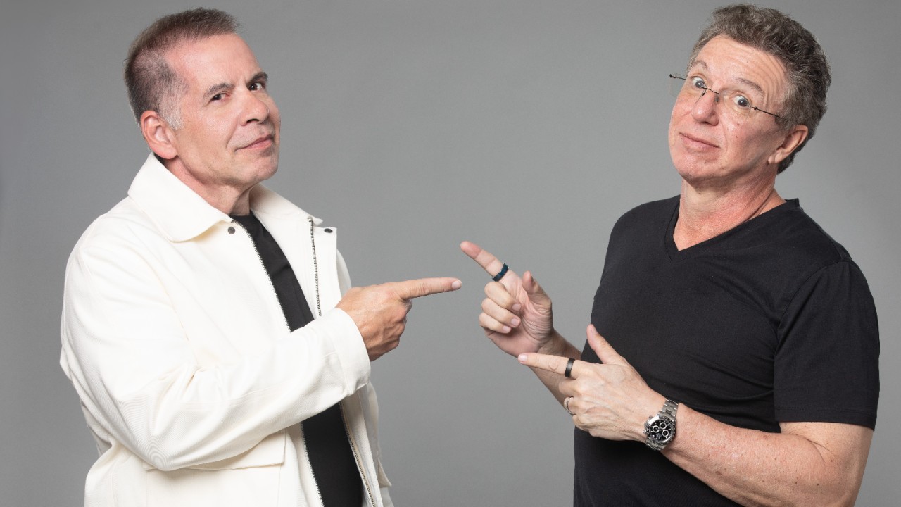 Leandro Hassum e Boninho apontam um para o outro em foto promocional com fundo neutro