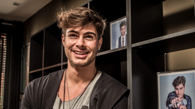 Léo Régis interpretado por Rafael Vitti sorri em ambiente interno com estante e fotos ao fundo em cena de Rock Story