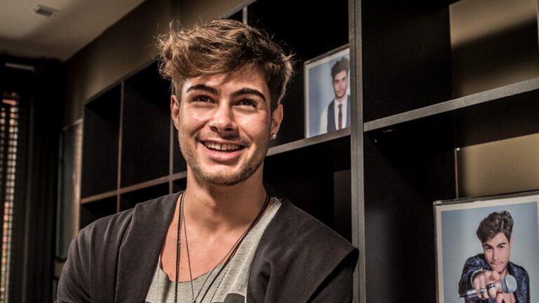 Léo Régis interpretado por Rafael Vitti sorri em ambiente interno com estante e fotos ao fundo em cena de Rock Story