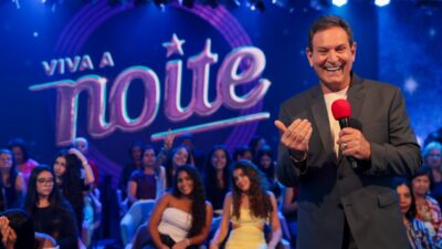 Luis Ricardo sorri com microfone vermelho no palco do Viva a Noite com plateia ao fundo no SBT