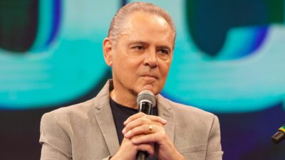 Luis Roberto segura microfone e fala em estúdio de TV com fundo iluminado em tons de azul