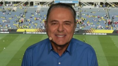 Luis Roberto sorri com microfone de lapela durante transmissão em estádio de futebol com arquibancadas e campo ao fundo