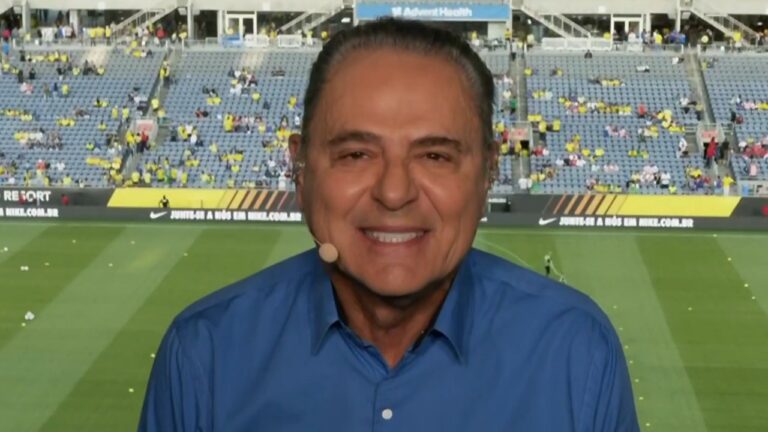 Luis Roberto sorri com microfone de lapela durante transmissão em estádio de futebol com arquibancadas e campo ao fundo