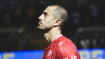 Marquinhos Pedroso de perfil com camisa vermelha de time de futebol durante partida em campo com arquibancada desfocada ao fundo