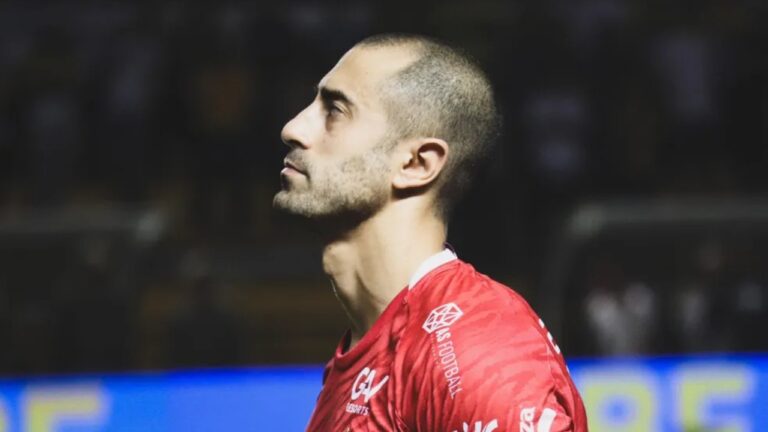 Marquinhos Pedroso de perfil com camisa vermelha de time de futebol durante partida em campo com arquibancada desfocada ao fundo
