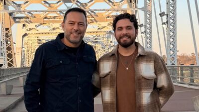 Maurício Ferraz e Gustavo Mioto sorrindo lado a lado em ponte metálica com estrutura de ferro ao fundo ao ar livre