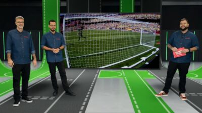 Mauro Beting, Pedro Oliveira e João Sales no estúdio do Xsports diante de telão com lance de futebol próximo ao gol e arquibancada ao fundo