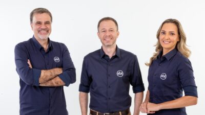 Mauro Beting, Tiago Leifert e Nadine Bastos posam sorrindo com camisas azul-marinho com logo do SBT em foto promocional de transmissão esportiva
