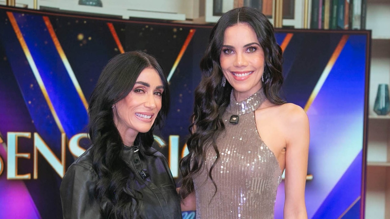 Michelle Barros e Daniela Albuquerque sorriem lado a lado em estúdio do Sensacional com telão iluminado ao fundo