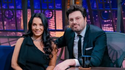 Mônica Carvalho e Danilo Gentili sentados lado a lado no The Noite sorrindo no estúdio com cenário urbano iluminado ao fundo