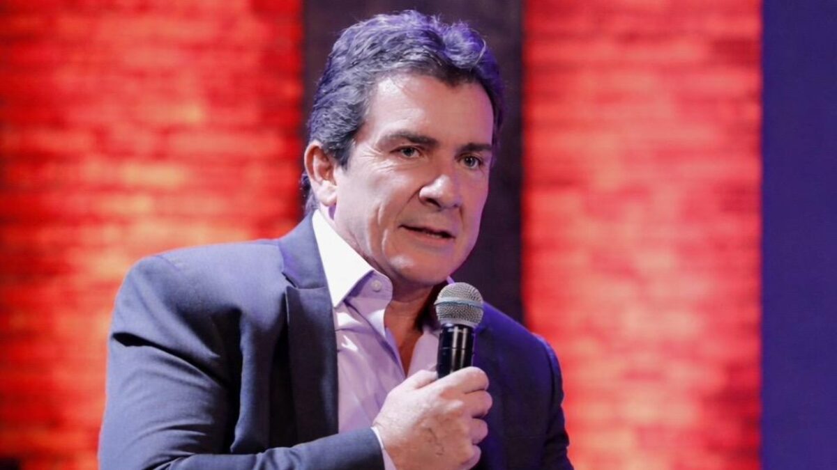 Murilo Fraga segura microfone e fala em evento com iluminação vermelha ao fundo vestindo terno escuro e camisa clara