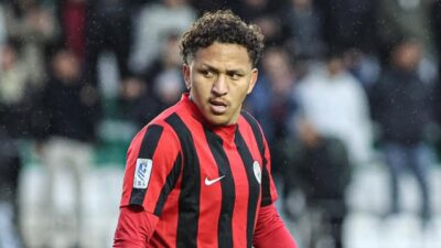 Jogador Neto Acará com camisa listrada em vermelho e preto da Nike em campo durante partida de futebol com fundo desfocado