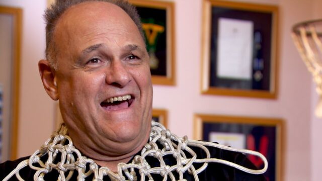 Oscar Schmidt sorri usando rede de basquete no pescoço em ambiente com quadros e troféu ao fundo