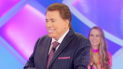 Silvio Santos sorrindo de terno e gravata durante gravação no palco do SBT com cenário colorido ao fundo