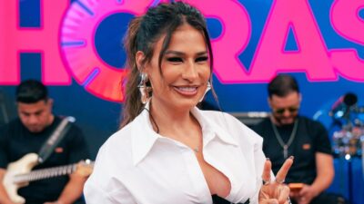 Simone Mendes sorri e faz gesto com a mão no palco do Altas Horas com banda ao fundo e iluminação colorida