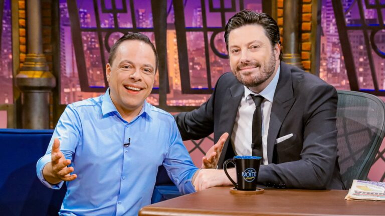 Thiago Gardinali e Danilo Gentili sorriem no palco do The Noite sentados à mesa com caneca do programa e cenário urbano ao fundo