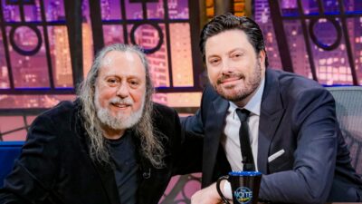Caio Fábio e Danilo Gentili sorrindo durante entrevista no programa The Noite com cenário iluminado ao fundo