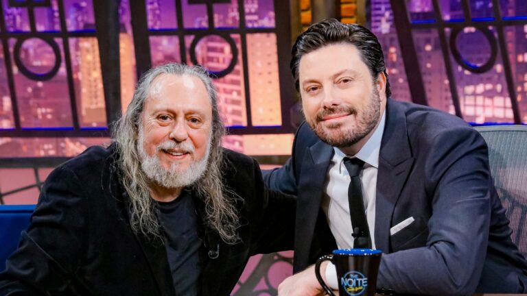 Caio Fábio e Danilo Gentili sorrindo durante entrevista no programa The Noite com cenário iluminado ao fundo