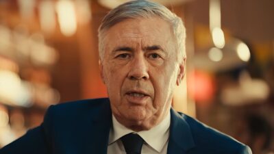 Carlo Ancelotti de terno e gravata durante comercial da Brahma em ambiente com iluminação quente ao fundo