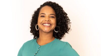 Denise Thomaz Bastos sorri usando blusa verde e brincos de argola em retrato com fundo claro