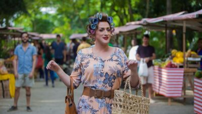 Paulo Gustavo como Dona Hermínia caminha por feira ao ar livre com vestido floral e bobes no cabelo em Minha Mãe É Uma Peça 3