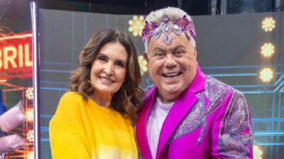Fátima Bernardes e Milton Cunha posam sorrindo nos bastidores de programa de TV com figurino rosa brilhante e tiara decorada