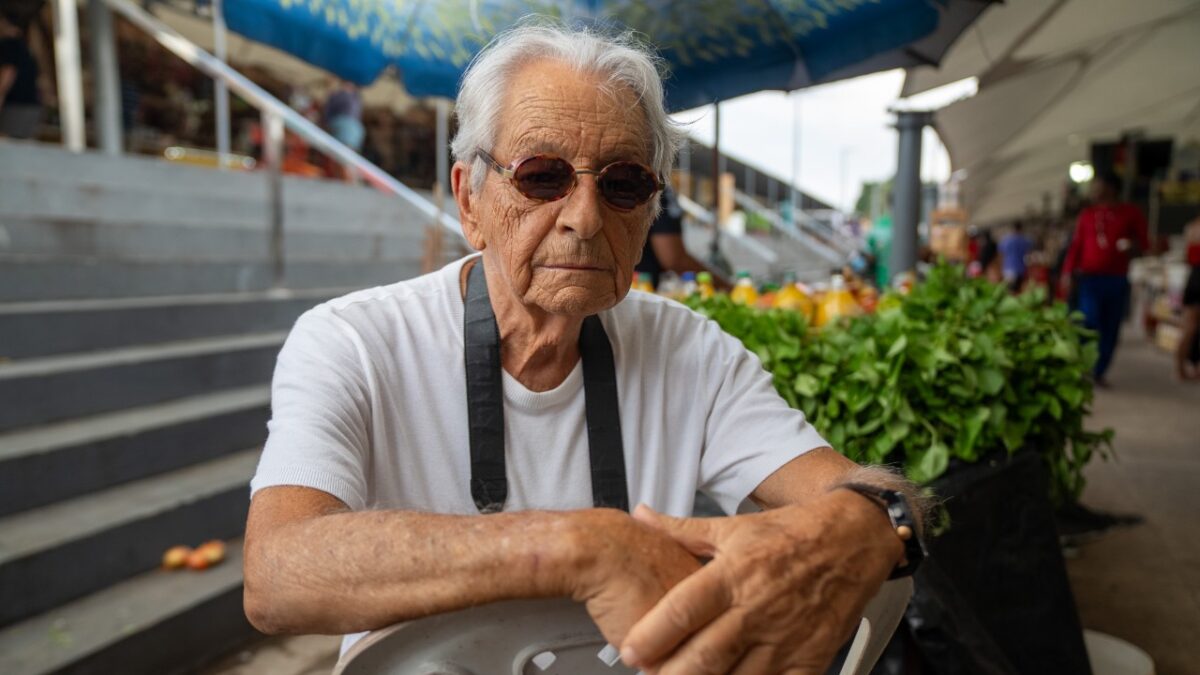 Fernando Gabeira sentado em feira ao ar livre usando óculos escuros e camiseta branca diante de barracas e escadaria