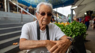 Fernando Gabeira sentado em feira ao ar livre usando óculos escuros e camiseta branca diante de barracas e escadaria