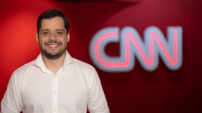 Fernando Miranda sorri em frente ao logotipo da CNN usando camisa branca em ambiente com fundo vermelho