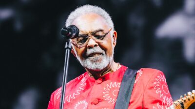 Gilberto Gil canta no palco com camisa vermelha estampada, óculos escuros e guitarra durante apresentação musical