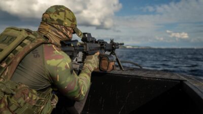Militar camuflado aponta metralhadora em embarcação durante operação marítima em alto-mar sob céu nublado