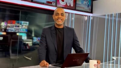 Jairo Nascimento sorri no estúdio da CNN Brasil diante de bancada com notebook e caneca da emissora