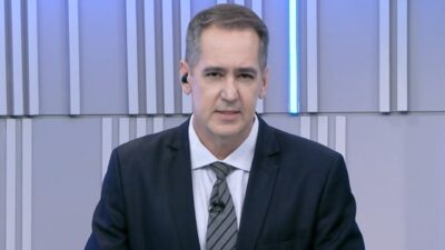 Luciano Cabral apresenta o JA2 da TV Anhanguera usando terno escuro e gravata em estúdio com painéis cinza ao fundo