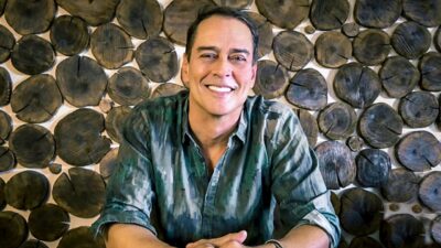 Marcello Antony sorri diante de parede com discos de madeira durante gravação em ambiente interno