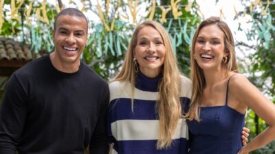 Thiago Oliveira, Maria Beltrão e Talitha Morete sorriem juntos em área externa decorada com fitas coloridas ao fundo
