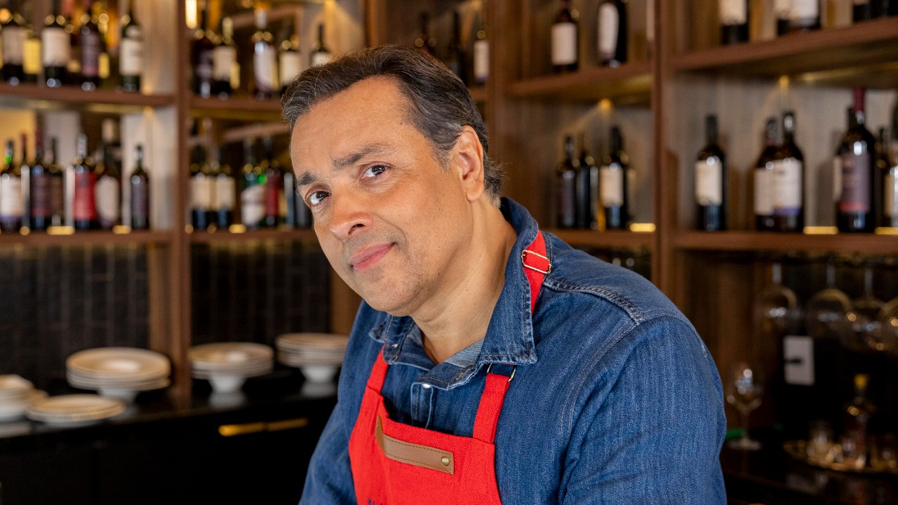 Nico (Rodrigo Fagundes) usa avental vermelho diante de adega com garrafas de vinho em cena de Quem Ama Cuida